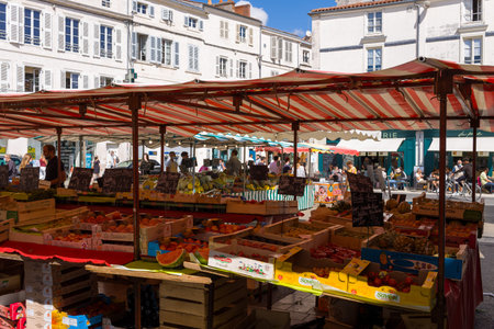 La Rochelle, France - August 31, 2020: People shop at outdoor market stalls selling fruits and vegetables under striped canopies in a city square with historic residential buildings and cafes in the background.のeditorial素材