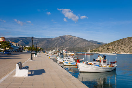 Port de Kilada, Greece - November 23, 2020: Fishing boats are moored along the quay beside a waterfront street lined with benches, palm trees, and buildings, with hills and wind turbines visible in the background under a clear blue sky.のeditorial素材
