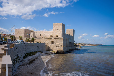 Trani, Italy - October 16, 2020: The historic stone Castello Svevo with towers and battlements is visible on the waterfront, with flags and parked cars nearby under a partly cloudy sky.のeditorial素材