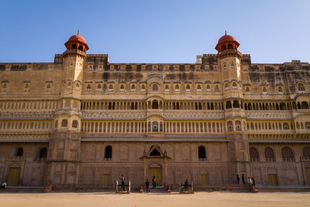 Bikaner, India - March 31, 2022: The front facade of the Junagarh Fort palace complex is shown under clear blue sky, featuring ornate sandstone architecture, arched windows, balconies, and people walking in the courtyard.のeditorial素材