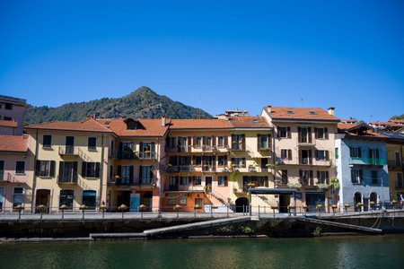 Ville de Omegna, Italy - September 28, 2020: Multi-story residential buildings with balconies and red-tiled roofs line the waterfront, with a mountain and clear blue sky in the background.のeditorial素材