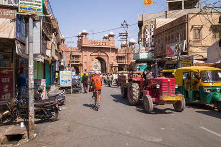 Bikaner, India - March 31, 2022: A busy street scene shows a historic arched gate in the background, with motorcycles, a red tractor, auto rickshaws, cyclists, pedestrians, and shops lining both sides of the road under a clear sky.のeditorial素材