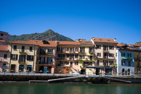 Ville de Omegna, Italy - September 28, 2020: Multi-story residential buildings with balconies and red-tiled roofs line the waterfront, with a mountain and clear blue sky in the background.のeditorial素材