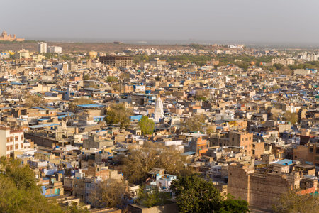 Jodhpur, India - March 26, 2022: An elevated view shows a densely packed urban area with blue-painted houses, flat rooftops, trees, and a white temple visible among the buildings under a hazy sky.のeditorial素材