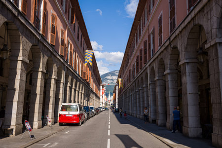 Chambery, France - September 23, 2020: A straight street lined with historic arcaded buildings on both sides, with parked cars, pedestrians, and a mountain visible in the background under a blue sky.のeditorial素材