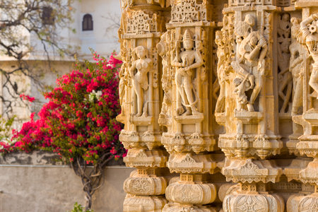 Chittorgarh, India - March 21, 2022: The image shows detailed stone carvings of human figures and ornate patterns on the columns of the Jain Shvetambar Temple, with a flowering bush and part of a white building visible in the background.のeditorial素材