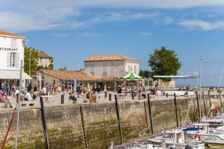 Ile de Re, France - September 1, 2020: People are walking, sitting, and socializing along a stone harbor promenade with moored boats, a restaurant, and a green-and-white striped canopy visible under a partly cloudy sky.のeditorial素材