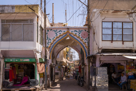 Jodhpur, India - March 26, 2022: A decorated arched gateway stands between two buildings with shops on either side, with people visible inside the shops and pedestrians further down the narrow street under overhead wires.のeditorial素材