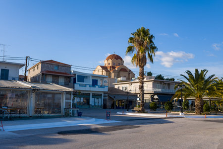 Port de Kilada, Greece - November 23, 2020: A street scene shows palm trees, local businesses, residential buildings, and a large domed church in the background under a clear blue sky.のeditorial素材