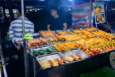 Can Tho, Vietnam - January 19, 2020: A variety of grilled skewers, sausages, and vegetables are displayed on metal trays at a night market food stall, with two men standing behind the counter under artificial lighting.のeditorial素材