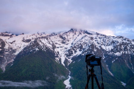 Unknown location - June 9, 2021: A digital camera mounted on a tripod is positioned in the foreground, facing towards a backdrop of snow-covered mountain peaks and forested slopes, likely in the Alps.のeditorial素材