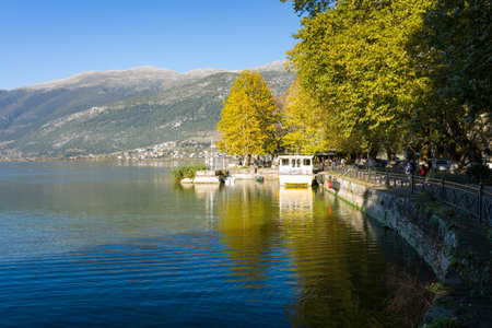 Ioannina, Greece - October 22, 2020: A lakeside promenade along Lake Ioannina features several moored boats, a tree-lined walkway with autumn foliage, and distant mountains under a clear blue sky.のeditorial素材