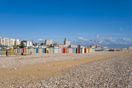 Le Havre, France - August 25, 2020: Rows of beach huts with vertical colored stripes stand on a pebble and sand beach, with people walking and sitting, and modern apartment buildings and the tower of St. Josephs Church visible in the background under a clのeditorial素材