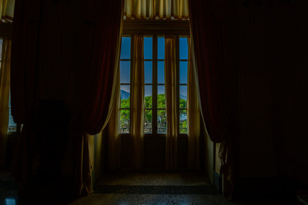 Isola Bella, Italy - September 30, 2020: The image shows a tall French window with yellow curtains in Palais Borromee, offering a view of green trees, distant mountains, and blue sky outside.のeditorial素材