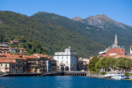 Ville de Omegna, Italy - September 28, 2020: Multi-story waterfront buildings with red tile roofs and balconies line the shore, with a white town hall building and a church spire visible against a backdrop of forested mountains under a clear blue sky.のeditorial素材