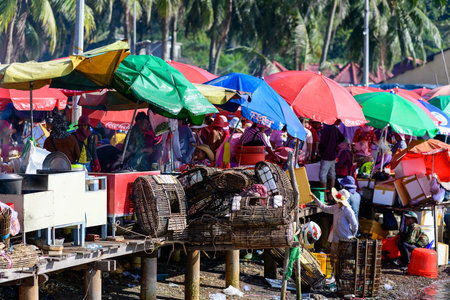 Marche de Kep, Cambodia - January 26, 2020: Vendors and customers are seen at outdoor seafood market stalls under red, green, blue, and yellow umbrellas, with woven baskets, coolers, and cooking equipment visible in a busy waterfront market scene.のeditorial素材