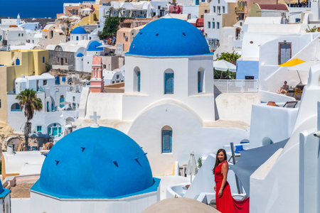 Oia, Santorini island, Greece - June 21, 2021: Beautiful girl in red dress and typical white-blue...