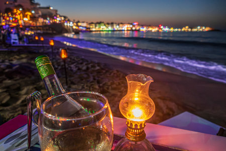 Hersonissos, Crete, Greece - July 27, 2021: Beautiful evening in Hersonissos bay. Romantic dinner served on the beach, with a bottle of wine and light of a one retro oil lamp.のeditorial素材
