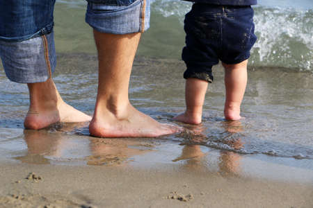 Feet, big and very small,  of the men and baby, father and son, standing in shallow water waiting for the wave, learning, playing and growing up togetherの写真素材
