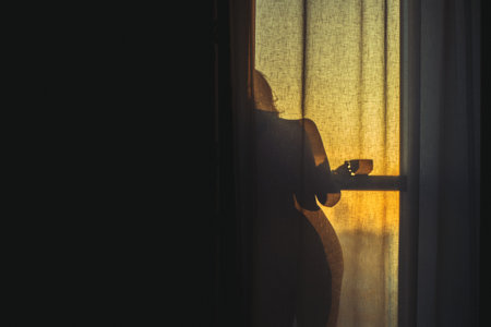 A naked girl is standing by the window behind the curtain and drinking coffee at dawnの写真素材