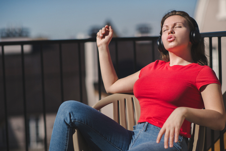 Beautiful girl in a red t-shirt and headphones. A girl on the balcony listening to music and singingの写真素材
