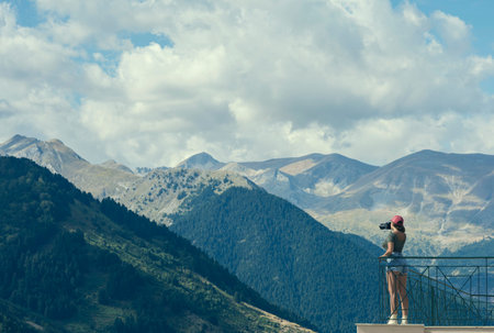A female photographer in a red cap with a camera stands on the balcony opposite greek mountains and...