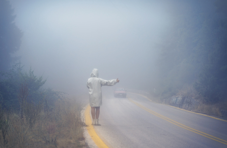 Young female in raincoat on the road in the fog. Travel of women in the raincoat hitchhiking in the rainの写真素材