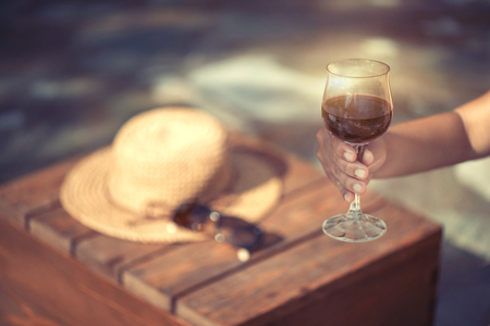 Glass of red wine in female hand. Sunglasses and thatch hat on the wooden tableの写真素材