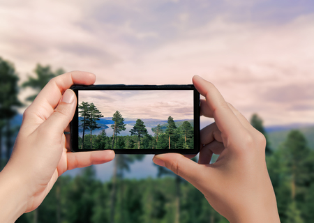 Picture of forest in Norway on mobile phone. Female take picture trees on mountain and lake background on smart phone.の写真素材