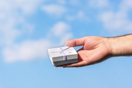 Male hand with a small white gift boxe on the blue sky background. Concept.の写真素材
