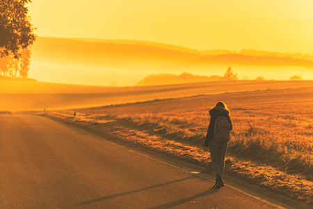 Young woman in the winter jacket with a backpack on the road against the background of the sunrise autumn field. Travel and tourism conceptの写真素材