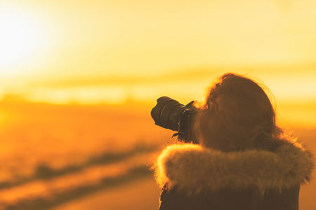 Young woman in the winter jacket with camera on the road against the background of the sunrise autumn field. Travel and tourism conceptの写真素材
