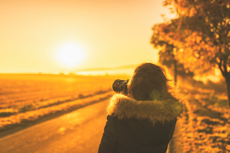 Young woman in the winter jacket with camera on the road against the background of the sunrise autumn field. Travel and tourism conceptの写真素材