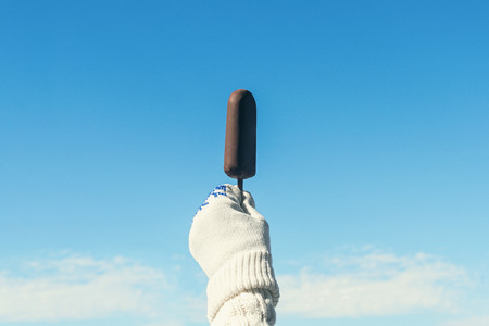 Female hand in knitted gloves with chocolate ice cream on the stick in the winter snowfall background. Conceptの写真素材