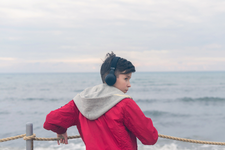 A boy in a red jacket and headphones looks at the sea waves in stormy rainy weatherの写真素材