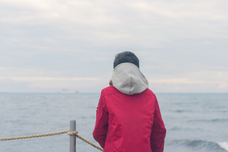 A boy in a red jacket and headphones looks at the sea waves in stormy rainy weatherの写真素材