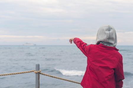 A boy in a red jacket and headphones looks at the sea waves in stormy rainy weatherの写真素材