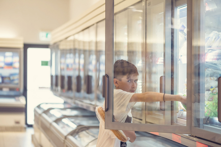 Boy in the supermarket opens glass doors of the freezer and trying to steal foodの写真素材