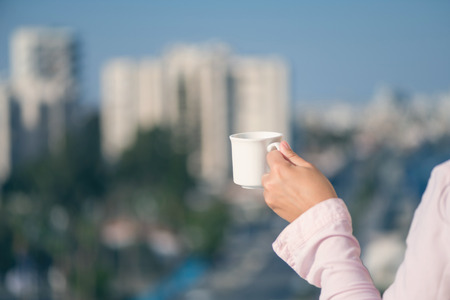 Woman hand in a pink shirt with cup of coffee at the summer morning against the background of resort townの写真素材