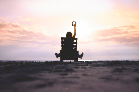 Girl in headphones is sitting on the sun lounger listening to music on the beach at sunset. Summer concept.の写真素材
