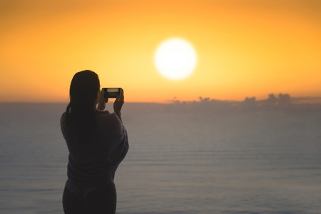 Silhouette of woman with wet hair wrapped in a blanket after swimming. Female taking picture on mobile phone standing on the beach opposite sunset sea.の写真素材