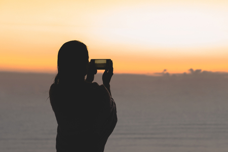 Silhouette of woman with wet hair wrapped in a blanket after swimming. Female taking picture on mobile phone standing on the beach opposite sunset sea.の写真素材