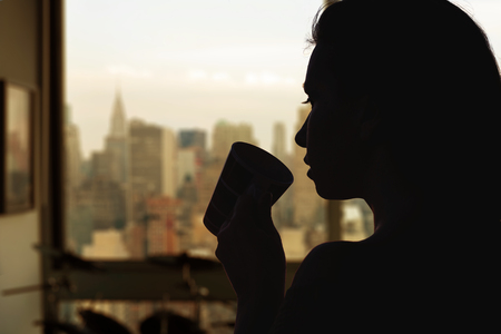 Silhouette of woman with cup of tea in the apartment with New York city view.の写真素材