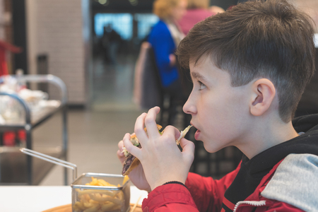 Boy eating fast food in the cafe. The concept of food and unhealthy lifestyle. French fries in basket and big hamburger with ketchup and sauceの写真素材