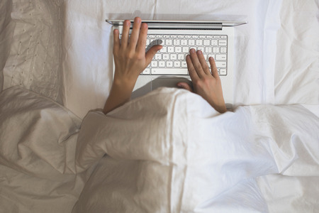 Girl lying with a laptop in bed covered by blanket. Woman can't sleep and have to work late at night. Top viewの写真素材