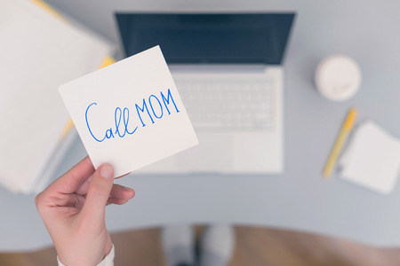 Woman clerk is sitting at office table holding note sticker with message "call mom" and going to call up her mother.の写真素材