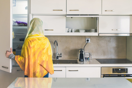 Back view on a muslim woman in a scarf opening door of a fridge on the kitchen. Preparation of breakfastの写真素材