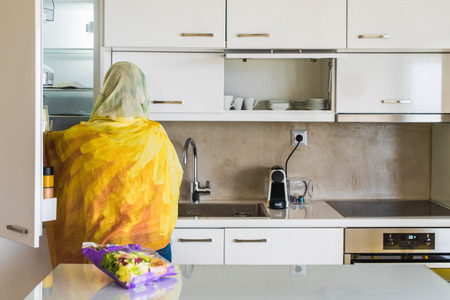 Back view on a muslim woman in a scarf opening door of a fridge on the kitchen. Preparation of breakfastの写真素材