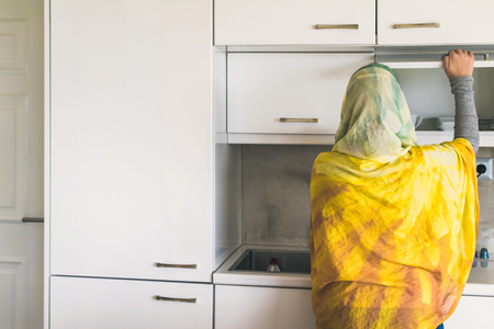 Back view on a muslim woman in a scarf opens door of a whte cabinet with cups and platesの写真素材