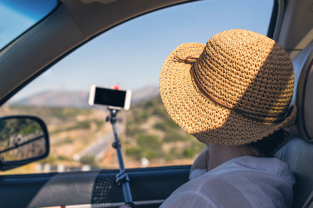 Blogger girl in the straw hat taking selfie picture or video using smartphone and selfie stick inside of car. Holidays and tourism conceptの写真素材